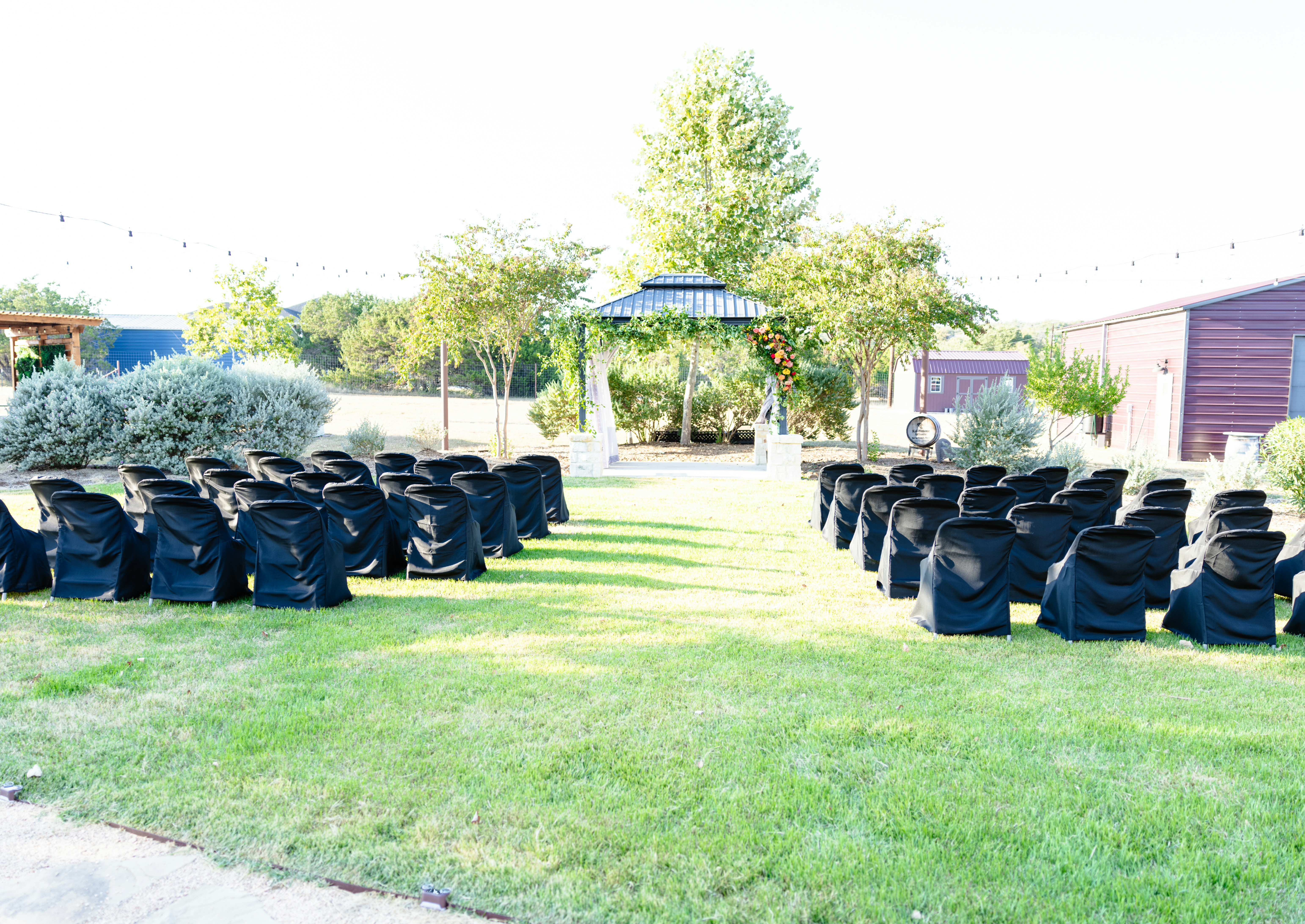 Outdoor wedding ceremony setup with black chair covers, floral arch gazebo, string lights and lush green lawn at Kai-Simone Winery